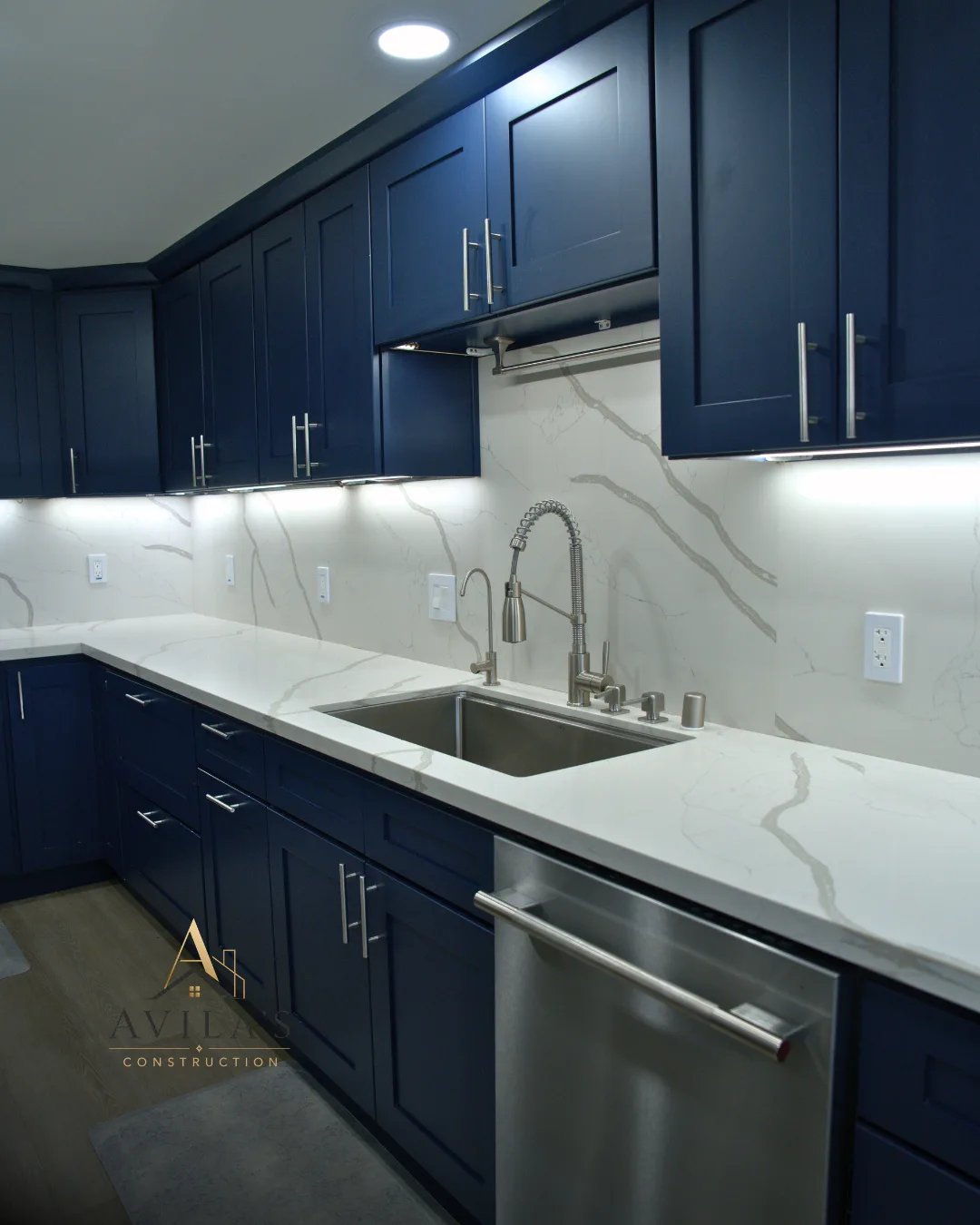 Finished kitchen remodel with deep navy cabinets, quartz waterfall counters and a marble backsplash, with the Avila's Construction logo in the lower left.